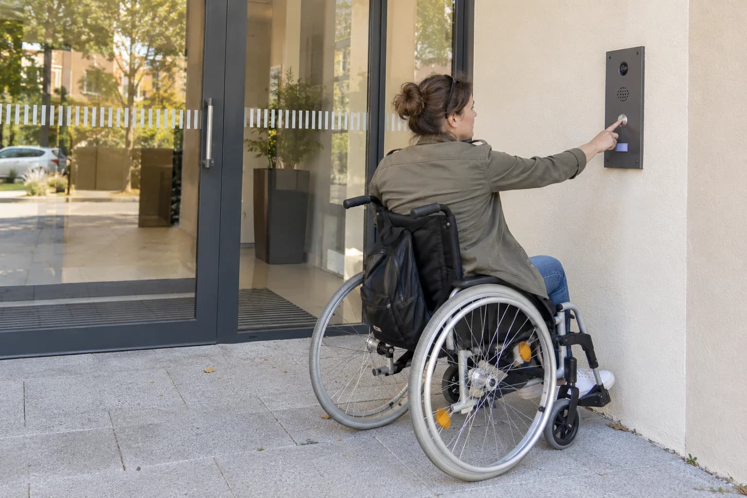 Illustration générée : une visiteuse en fauteuil utilise le bouton d’un interphone générique devant une entrée fictive